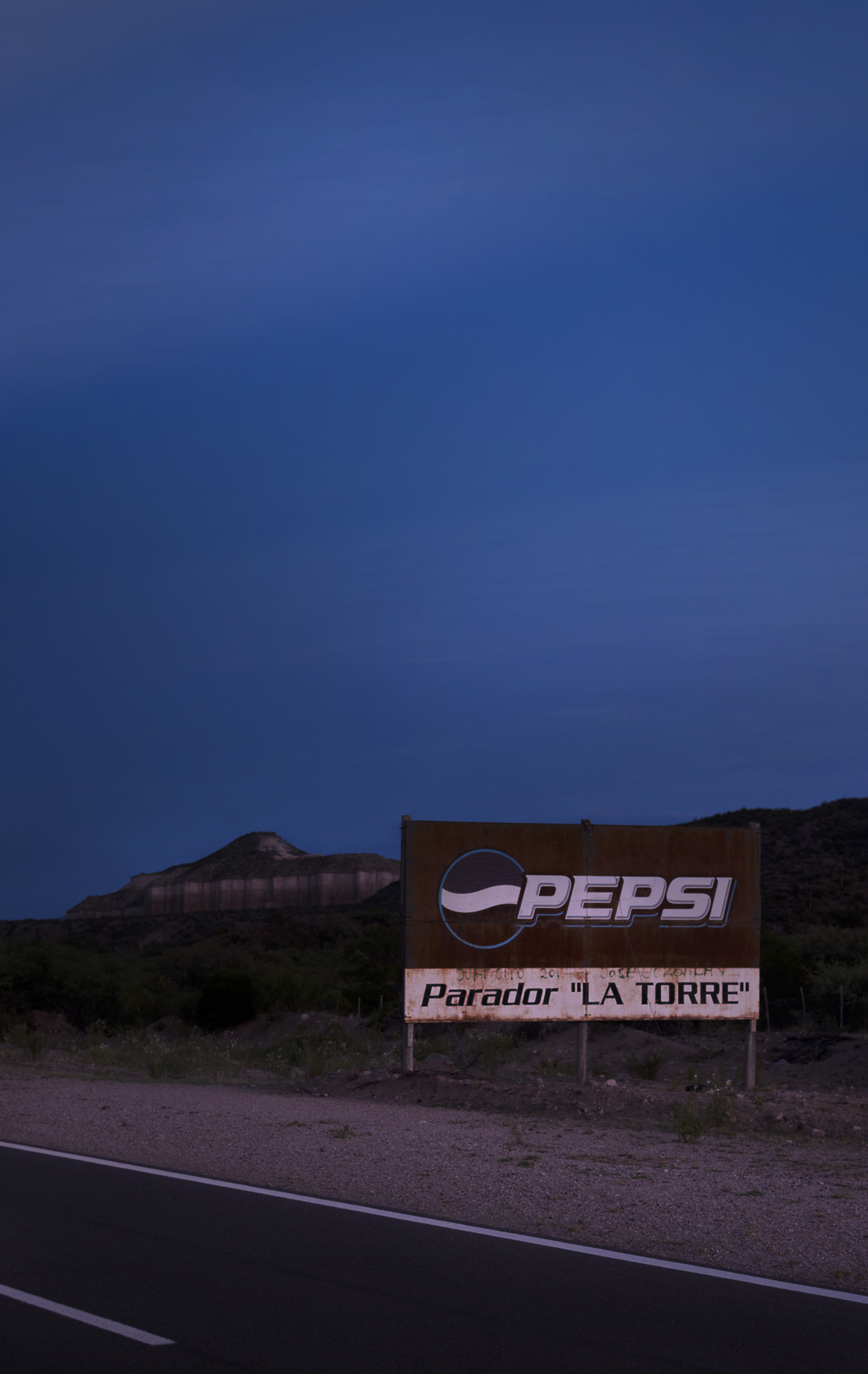 A rusty Pepsi billboard stands against a peaceful desert landscape with clear blue skies in the afternoon. The Pepsi billboard says "Parador la Torre", spanish for "La Torre Gas Station".