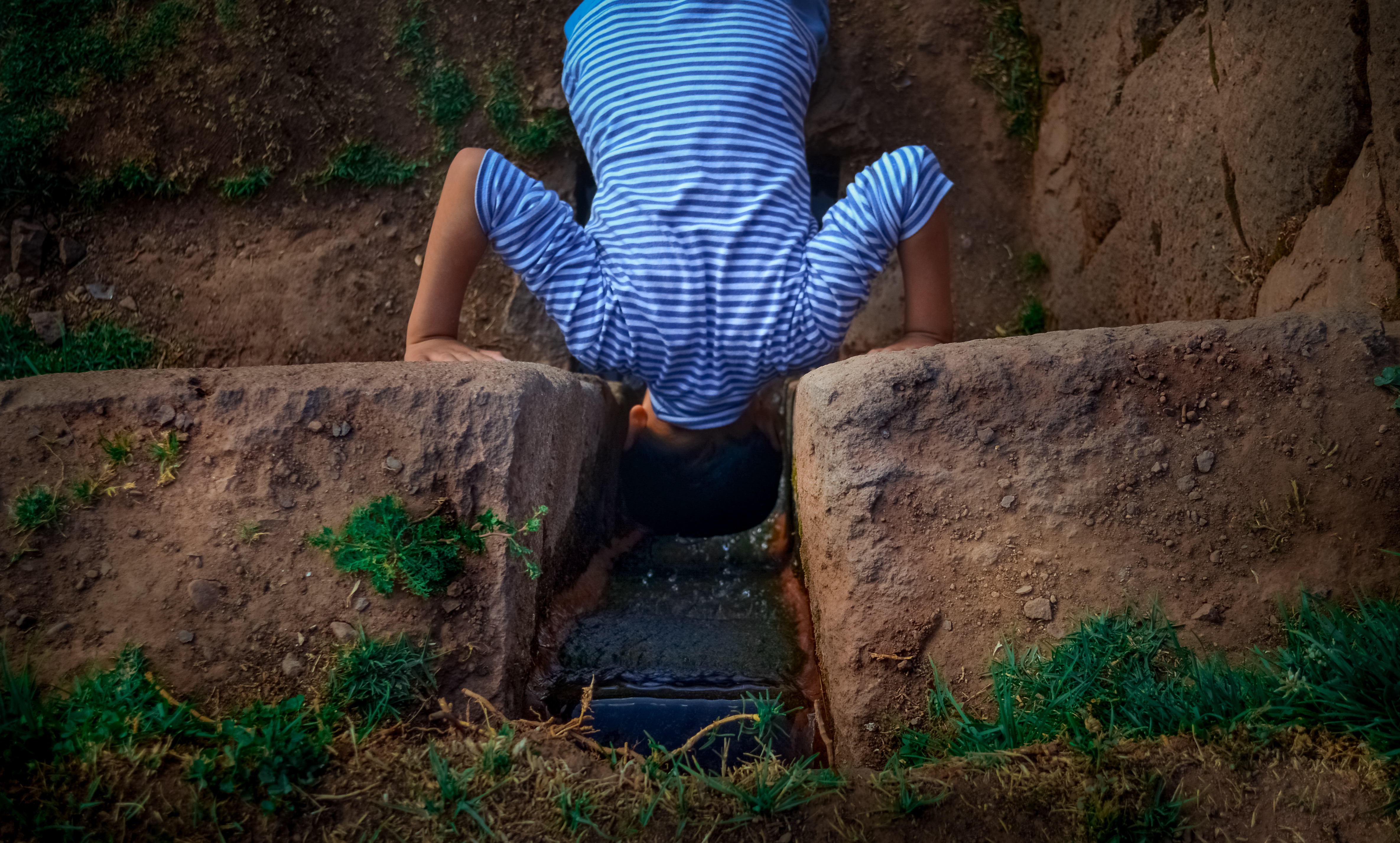 A small boy wearing a light blue striped shirt dips is head into a water stream carved out of a rock wall