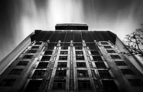 A black and white picture of a tall symmetrical building of international style. The building is covered by a thin veil and a layer of graffiti. White clouds drift across the dark sky above the building.