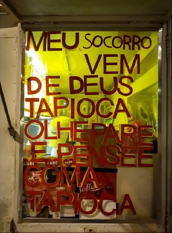A window in a food truck with a red sign saying "MEU SOCORRO VEM DE DEUS TAPIOCA OLHE PARE E PENSE COMA TAPIOCA", portuguese for "MY SALVATION COMES FROM GOD TAPIOCA LOOK STOP AND THINK EAT TAPIOCA". Behind the window we can see the food truck's kitchen illuminated by bright yellow lights and a man wearing white cloths and a hat.