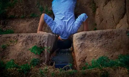 A small boy wearing a light blue striped shirt dips is head into a water stream carved out of a rock wall
