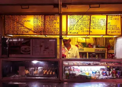 A food truck's counter illuminated in a dim red light. On top of the counter there is a bright yellow sign listing multiple flavors of tapioca. Inside the truck we can see a man in white cloths working.
